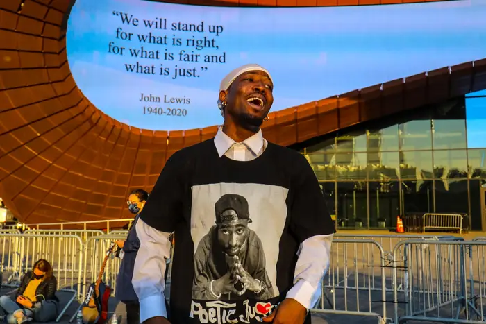 A Black man, wearing a shirt of Tupac Shakur, smiles while in front of a quote from John Lewis—"We will stand up for what is right, what is fair and what is just"—that is projected on the Barclays Center